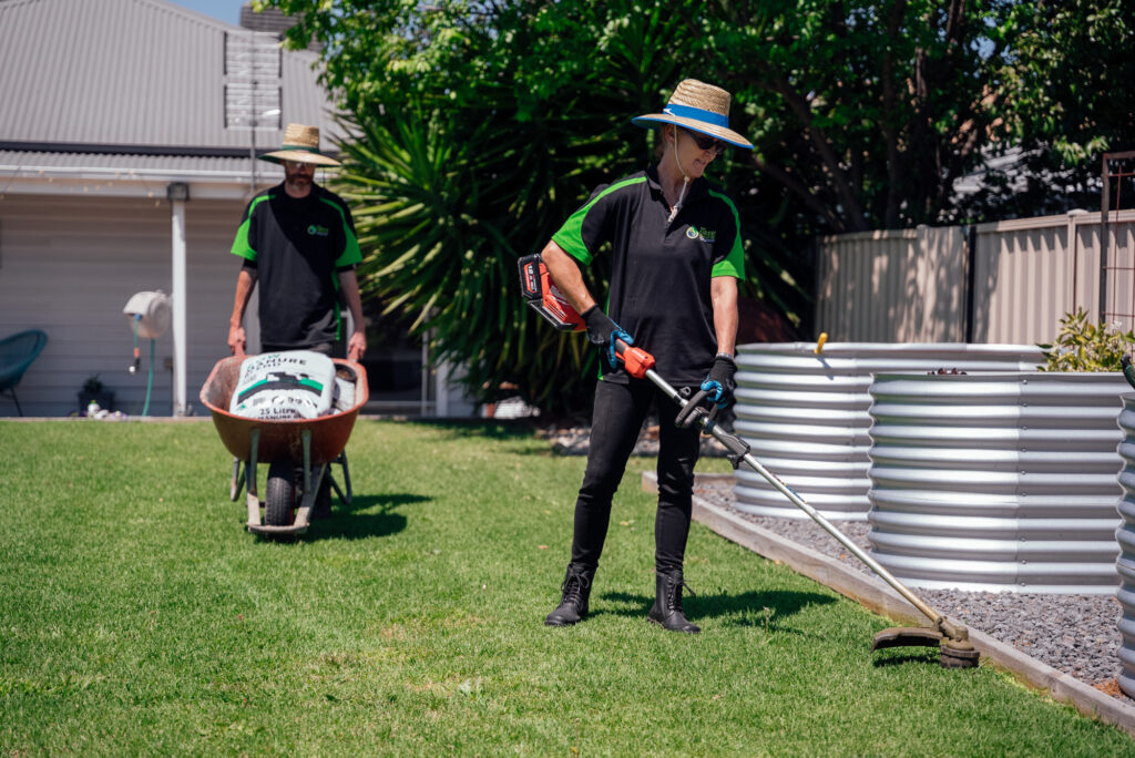 Two gardeners working in a residential yard, one trimming lawn edges with an electric whipper snipper and the other pushing a wheelbarrow. The Green Clean Team providing eco-friendly gardening and outdoor maintenance across Bendigo, Castlemaine, Echuca, and Central Victoria