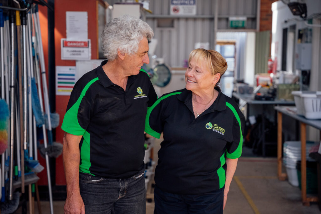 Green Clean Team co-founders, Mike and Sandra, stand in a warehouse surrounded by various cleaning supplies on shelves