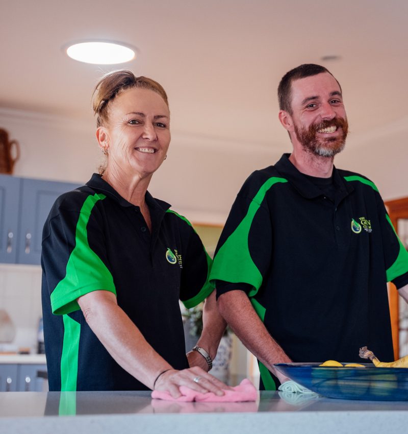 Two people in green and black shirts stand at a counter. They are smiling and the person on the left is cleaning.